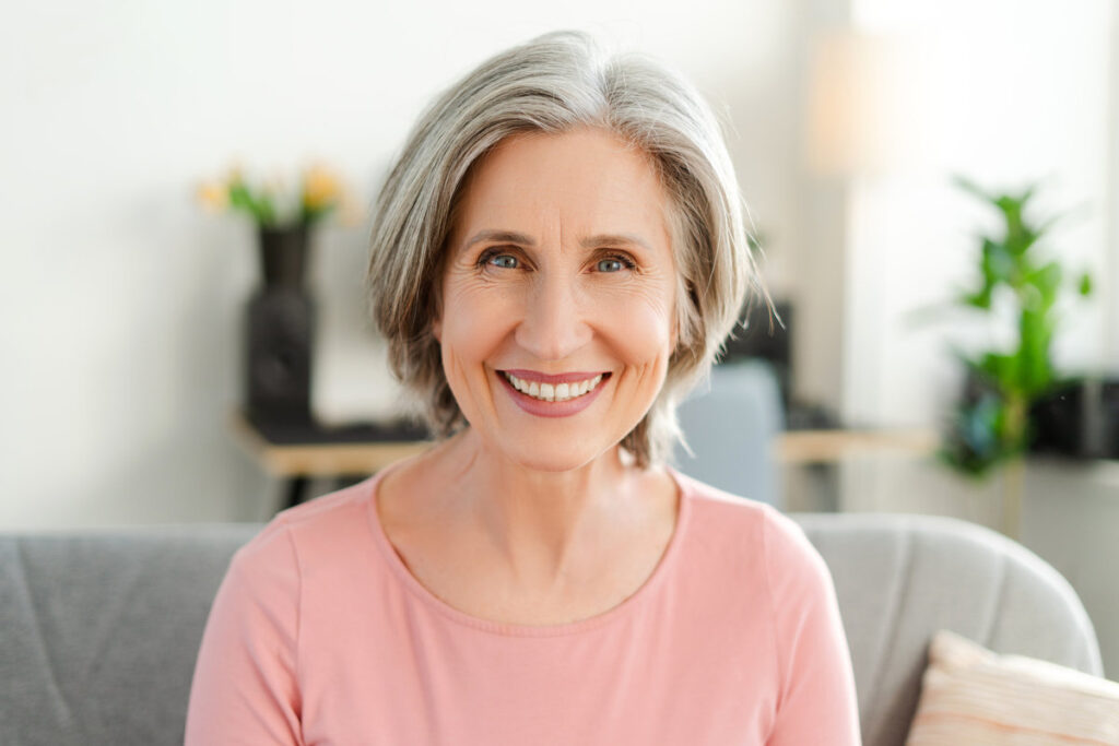 Confident smiling senior woman sitting on sofa looking at camera. Portrait happy retired female with stylish gray hair, white teeth at home. Natural beauty, healthy lifestyle concept