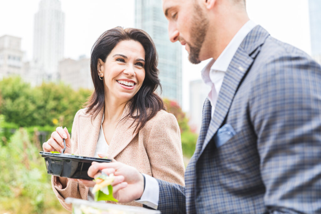 Business people having lunch at park in Chicago. Man and woman, wearing smart casual clothes, eating pasta and salad on lunch break. Downtown skyscrapers on background
