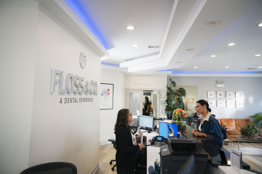 Floss & Co dental office reception area featuring a staff member at the front desk assisting a patient, with the branded "Floss & Co – A Dental Experience" logo on the wall, blue ambient lighting along the ceiling, and a modern, welcoming waiting area in the background.