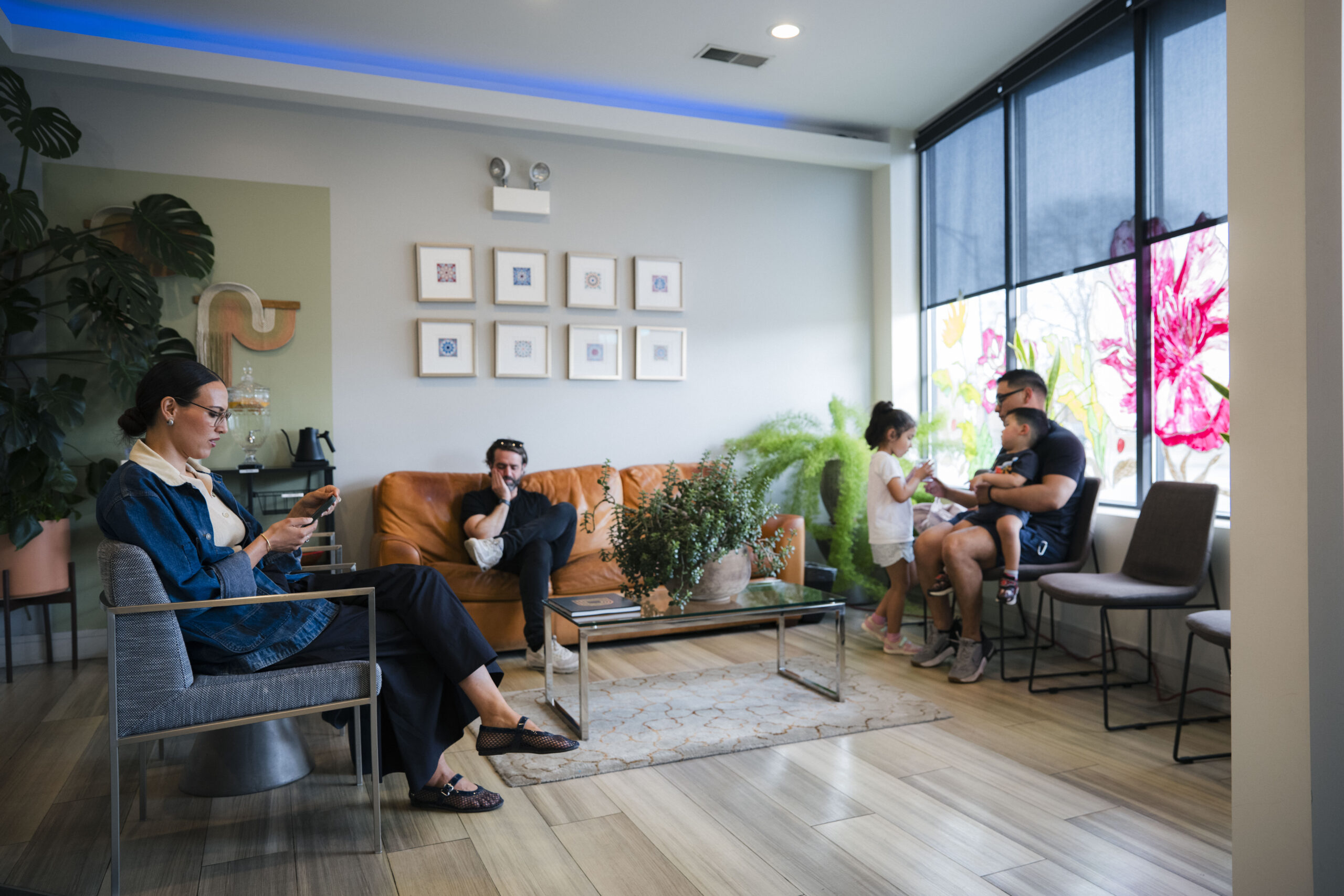 Floss & Co. dental office waiting room with patients seated in modern lounge chairs and a leather sofa, surrounded by indoor plants, framed artwork, and large windows with floral decals